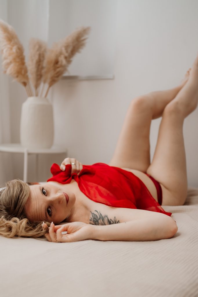 Boudoir Shooting: Frau liegt auf dem Bett in rotem Tüll, sanftes Tageslicht, intime Stimmung.